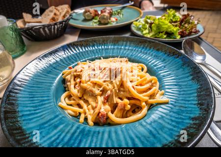 Spaghetti Carbonara auf blauem Teller, Salat, Brot, Getränk und Schweinebraten auf dem zweiten Teller auf dem Restauranttisch. Stockfoto