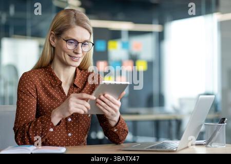 Eine Geschäftsfrau arbeitet an ihrem Schreibtisch, konzentriert sich auf ein Tablet, während sie lächelt, mit einem Laptop in der Nähe. Die Szene spielt in einem Büro. Stockfoto