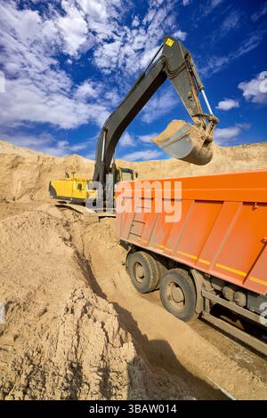 Gelber Bagger lädt Sand in einen Müllwagen in einem Bausteinbruch unter einem leuchtend blauen Himmel Stockfoto