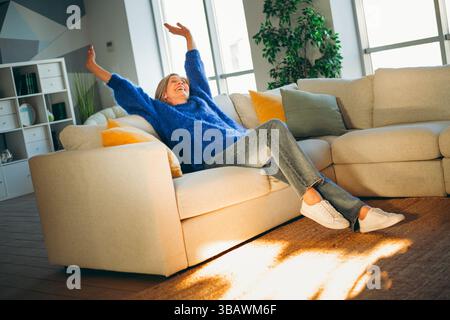Glückliche Frau, die sich tagsüber auf einem gemütlichen Sofa in einem hellen Wohnzimmer entspannt und einen ruhigen und erfrischenden Moment genießt Stockfoto
