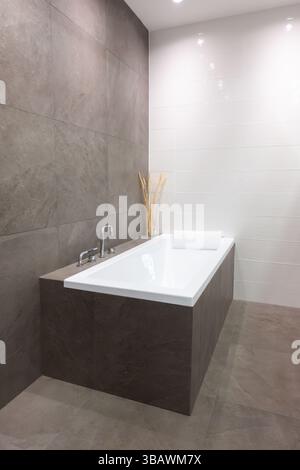 Moderne eingebaute Keramikbadewanne mit Edelstahl- und Chromarmaturen in einem modernen Badezimmer mit grauen Fliesenwänden und Boden für ein elegantes Zuhause Stockfoto