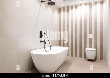 Elegantes und luxuriöses Badezimmer mit großen grauen Steinfliesen, freistehender Keramikbadewanne und moderner Toilette für modernes Innendesign Stockfoto