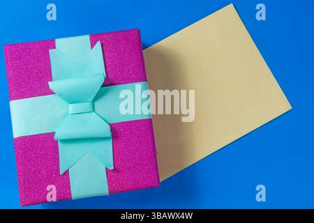 Geschenkbox mit rosa Glitzer und blauem Band und leerer Karte auf leuchtend blauer Oberfläche für Feiern, Geburtstage, Feiertage oder besondere Anlässe. Stockfoto