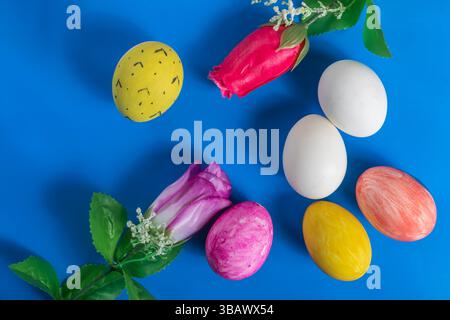 Farbenfrohe Ostereier und künstliche Blumen sind kreativ auf einem leuchtend blauen Hintergrund für die Osterferien angeordnet. Stockfoto