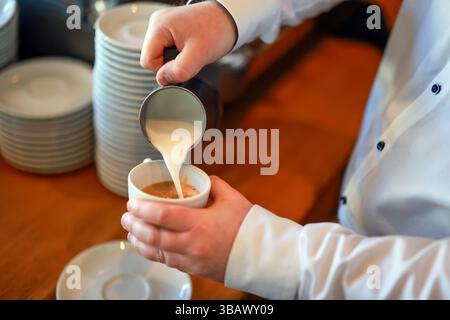 07.04.2025, Deutschland, - Barista bereitet einen Kaffee zu, hier einen Cappuccino, gießt Milchschaum aus einer kleinen Kaffeekanne von Hand in eine Tasse Espresso. 00X Stockfoto