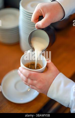 07.04.2025, Deutschland, - Barista bereitet einen Kaffee zu, hier einen Cappuccino, gießt Milchschaum aus einer kleinen Kaffeekanne von Hand in eine Tasse Espresso. 00X Stockfoto