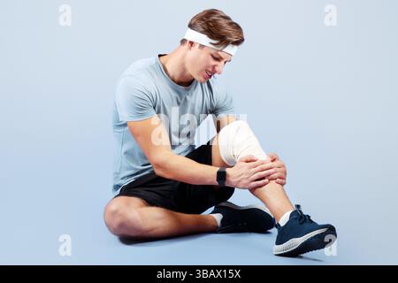 In voller Länge Porträt des jungen Sportlers mit Knieverletzung, hält seine verstauchten bandagierten Bein, violetten Studio-Hintergrund Stockfoto