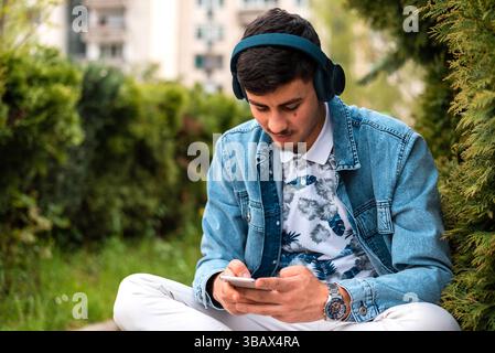 Ein junger glücklicher Mann mit Kopfhörern, der draußen ein Handy benutzt. Stockfoto