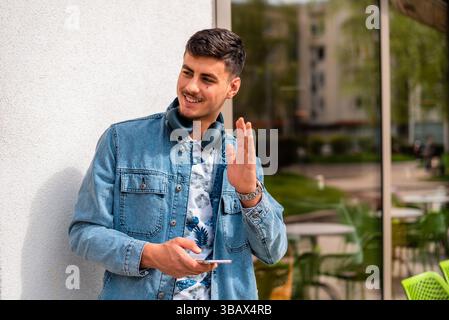 Ein junger glücklicher, stilvoller Mann, der ein Handy benutzt und mit der Hand einem Freund auf der Straße der Stadt zuwinkt. Stockfoto