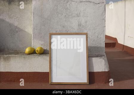 Leerer hölzerner Bilderrahmen, der sich an die strukturierte Wand auf dem Dach des spanischen Hauses lehnt. Zwei gelbe Zitronen auf einem Betonvorsprung. Natürliches Sonnenlicht, rustikal Stockfoto