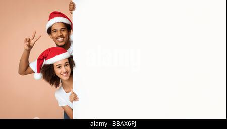Lächelndes junges afroamerikanisches Paar in Weihnachtsmützen, das herausguckt Von hinten weißes leeres Banner Stockfoto