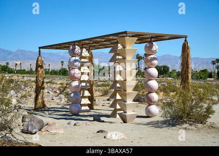 Palm Springs, Kalifornien, USA. Mai 2025. Die skulpturale Installation des Künstlers Kapwani Kiwanga für Desert X 2025, die Geometrie, Material und Landschaft zu einer meditativen Struktur verschmilzt, spielt in der felsigen Wüstenlandschaft in der Nähe von Palm Springs. Vier unterschiedliche Säulen, die jeweils aus gestapelten Formen wie Kugeln und Pyramiden aufgebaut sind, tragen ein Baldachin, das Schatten mit Muster wirft. An den Ecken hängen Bündel getrockneter Pflanzenfasern, die an indigene Architektur und natürliche Unterkünfte erinnern. Kiwangas Arbeit beschäftigt sich oft mit Macht, Widerstand und historischer Erinnerung. Hier spricht das Stück ruhig Stockfoto