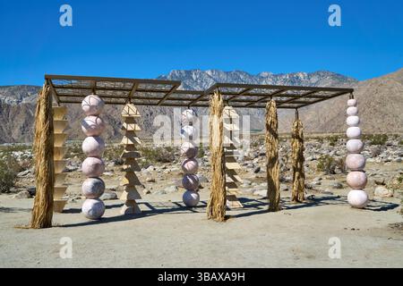 Palm Springs, Kalifornien, USA. Mai 2025. Die skulpturale Installation des Künstlers Kapwani Kiwanga für Desert X 2025, die Geometrie, Material und Landschaft zu einer meditativen Struktur verschmilzt, spielt in der felsigen Wüstenlandschaft in der Nähe von Palm Springs. Vier unterschiedliche Säulen, die jeweils aus gestapelten Formen wie Kugeln und Pyramiden aufgebaut sind, tragen ein Baldachin, das Schatten mit Muster wirft. An den Ecken hängen Bündel getrockneter Pflanzenfasern, die an indigene Architektur und natürliche Unterkünfte erinnern. Kiwangas Arbeit beschäftigt sich oft mit Macht, Widerstand und historischer Erinnerung. Hier spricht das Stück ruhig Stockfoto