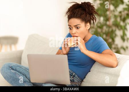 Black Lady Beißen Burger Mit Laptop Browsing Internet Sitting Indoor Stockfoto