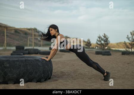 Fitnessbegeisterte nehmen an Liegestützübungen Teil, wobei sie einen großen Reifen als Stütze in einer rauen Outdoor-Umgebung verwenden. Stockfoto