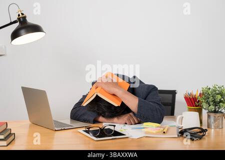 Ich arbeite spät im Büro. Gestresster junger Mann, der an einem Tisch zwischen Büchern sitzt. Überarbeiteter Kerl, der während seines Studiums im Stadtcafé schläft und Buch O schreibt Stockfoto