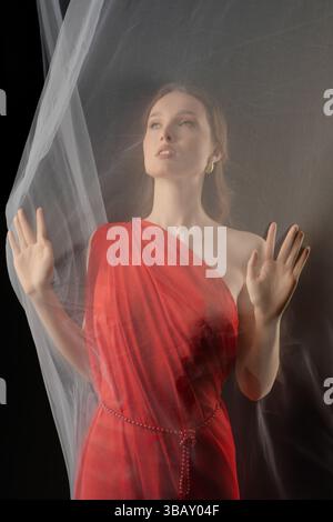 Frau posiert anmutig in einem roten Kleid mit transparentem Stoff in einem dramatischen Studio-Setting, das starke Emotionen weckt Stockfoto