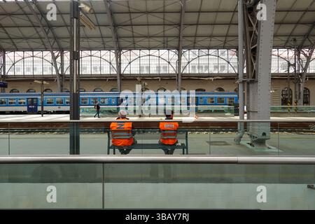 Zwei Eisenbahnarbeiter in orangefarbenen Westen sitzen auf einer Bank gegenüber den Gleisen, während ein Personenzug der Tschechischen Eisenbahn über den Bahnsteig in Prag Cent zu sehen ist Stockfoto