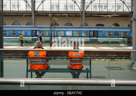 Zwei Eisenbahnarbeiter in orangefarbenen Westen sitzen auf einer Bank gegenüber den Gleisen, während ein Personenzug der Tschechischen Eisenbahn über den Bahnsteig in Prag Cent zu sehen ist Stockfoto