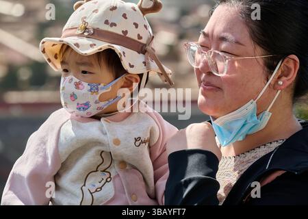 Mutter hält ein kleines, maskiertes Kind in gemusterter Kleidung und Hut, beide tragen Schutzmasken im Freien Stockfoto