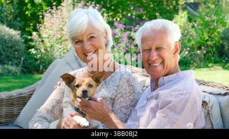 Porträt Eines Lächelnden Seniorenpaares Im Ruhestand, Das Auf Einem Außensitz Im Sommergarten Mit Haustierhund Sitzt Stockfoto