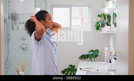 Frau Mit Schlafanzug, Die Vor Dem Badezimmerspiegel Steht Und Haare Hochlegt Stockfoto