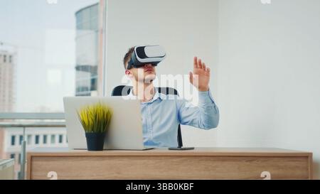 Mann mit VR-Headset im modernen Büro. Ein junger Mann, der an einem Schreibtisch in einem hellen Büro sitzt und ein Virtual-Reality-Headset trägt, während er mit dem interagiert Stockfoto