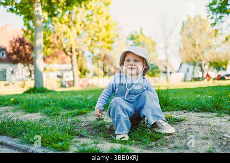 Ein glückliches Kleinkind, das im Park auf dem Gras sitzt, einen Sonnenhut trägt und die Natur genießt. Perfekt für Themen wie Kindheit, Freizeit und Sommerspaß Stockfoto