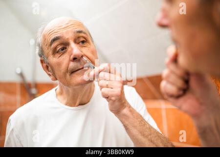 Porträt eines älteren Mannes, der Nasenhaare mit einer Pinzette entfernt, während er vor dem Badezimmerspiegel steht. Stockfoto