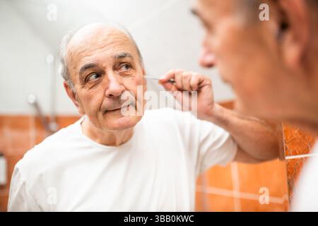 Porträt eines älteren Mannes, der Ohrhaare mit einer Pinzette entfernt, während er vor dem Badezimmerspiegel steht. Stockfoto