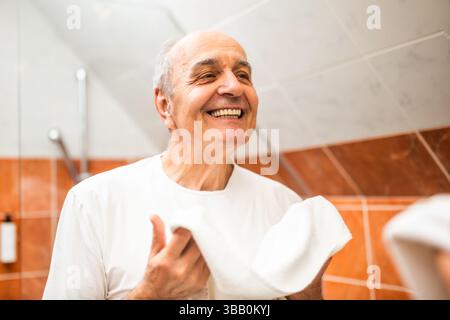 Zufriedener älterer Mann mit einem Handtuch in den Händen, der sein Spiegelbild im Spiegel überprüft und lächelt, nachdem er sich im Badezimmer Zähne und Gesicht gewaschen hat. Stockfoto