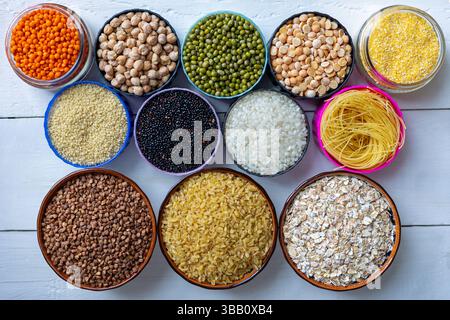 Draufsicht auf verschiedene Körner, Leguminosen und Pasta in Schüsseln auf weißem Holztisch. Verschiedene Körner, Hülsenfrüchte und Nudeln in bunten Schüsseln auf einem Stockfoto