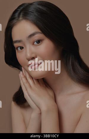 Schönes junges asiatisches Model mit perfekt sauberer und glatter Haut faltete ihre Hände in der Handfläche und legte es ihr ins Gesicht, steht auf einem Beige Stockfoto