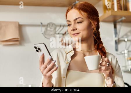 Die schöne rothaarige Frau schlürft ihren Kaffee aufmerksam, während sie ihr Smartphone zu Hause benutzt. Stockfoto