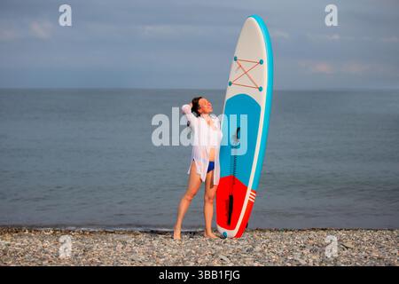 Eine Frau steht am Strand mit einem SUP-Board. Sie trägt ein weißes Hemd und blaue Badehosen. Stockfoto