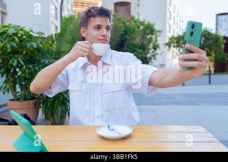 Junger Mann, der Selfie nimmt, während er draußen Kaffee trinkt, mit Tablet auf dem Tisch Stockfoto