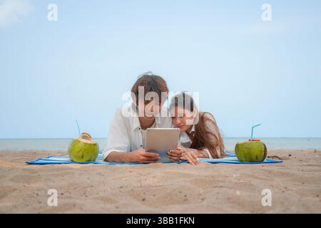 Junges asiatisches Paar, das am Strand liegt und einen entspannenden Moment genießt und Inhalte auf einem digitalen Tablet ansieht, mit Kokosnussgetränken auf jeder Seite. Ein gemütliches und Stockfoto