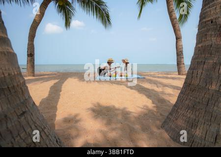Junges asiatisches Paar, das einen tropischen Strandmoment mit Gitarrenmusik und Kokosnussgetränken auf einer gestreiften Picknickmatte genießt. Die Frau klatscht mit, während der Mann Stockfoto