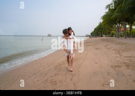 Junges asiatisches Paar verbringt schöne Zeit an einem tropischen Strand, mit dem Mann, der der Frau entlang der Küste einen Huckepack-Ritt gibt und Freude ausdrückt Stockfoto