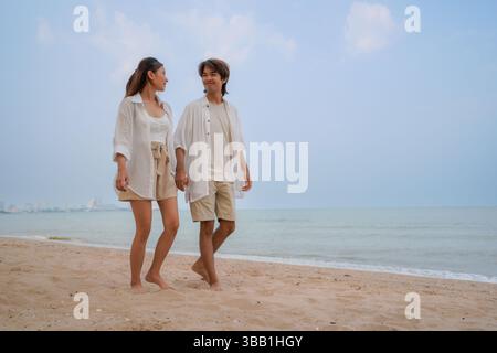 Junges asiatisches Paar, das einen entspannenden Spaziergang am Sandstrand unter klarem blauen Himmel genießt, gekleidet in passenden leichten, lässigen Outfits, die Romantik symbolisieren Stockfoto