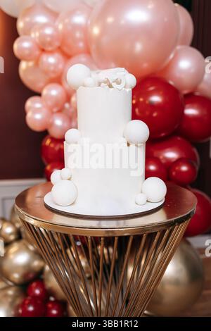 Weißer süßer Kuchen zur Geburtstagsfeier. Großer zweistöckiger Kuchen auf einem Hintergrund aus roten Ballons. Festlicher exquisiter Kuchen für das Geburtstagskind. Stockfoto