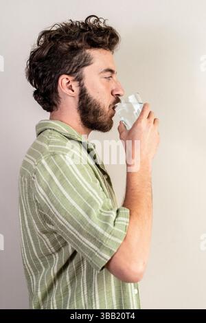 Seitenprofil-Porträt eines lächelnden Millennials in lässigem grünen Hemd, der klares Wasser aus einem Glas gegen eine neutrale Studiowand schlürft. Konzept von dai Stockfoto