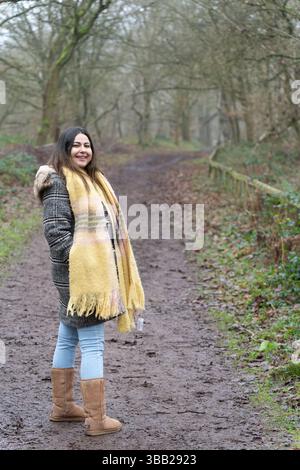 Lächelnde Frau in Mantel und Stiefeln, die im Winter im Sherwood Forest auf dem Waldweg steht Stockfoto
