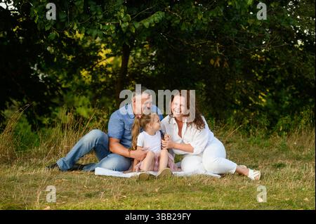 Familie, die Spaß hat, zusammen im Park zu sitzen, auf der Decke zu lachen und zu plaudern. Mom, Dad und Tochter genießen die Zeit draußen beim Picknick. Happy Family Weekend Konzept Stockfoto