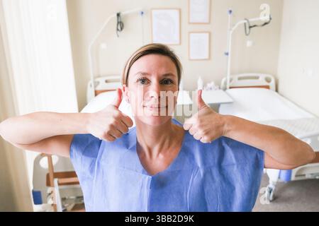 moskau, russland, 19.02.2025 Eine Frau im Krankenhauskleid gibt die Daumen hoch. Das Zimmer ist sauber und gut beleuchtet, mit einem Bett im Hintergrund. Die Frau i Stockfoto