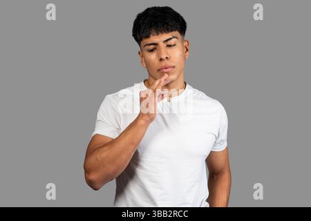 Nachdenklicher junger latino-Mann, der sein Kinn berührt, während er vor grauem Hintergrund denkt Stockfoto