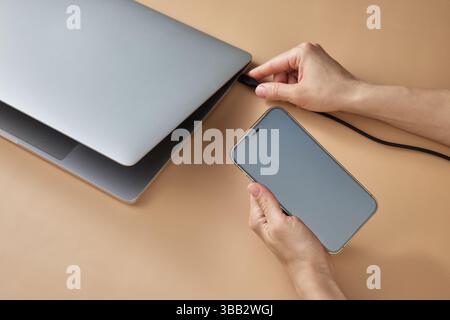 Weibliche Hand, die das Ladekabel mit dem Laptop verbindet und das Smartphone am beigefarbenen Tisch hält, Großansicht Stockfoto