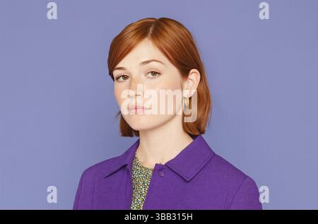 Eine junge Frau mit schulterlangen dunkelbraunen Haaren und Sommersprossen trägt ein lila Hemd mit Kragen vor einem massiven Lavendelhintergrund. Sie trägt einen goldenen Korb Stockfoto