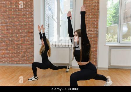 Zwei junge Frauen in schwarzer Sportswear, die mit erhobenen Armen in einem hellen Fitnessstudio mit Ziegelwänden und Holzboden Ausfallhosen. Stockfoto