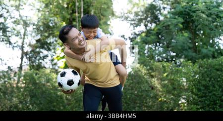 Aktivität: Vater trägt den Sohn auf dem Rücken, während er im Garten einen Fußball hält. Stockfoto
